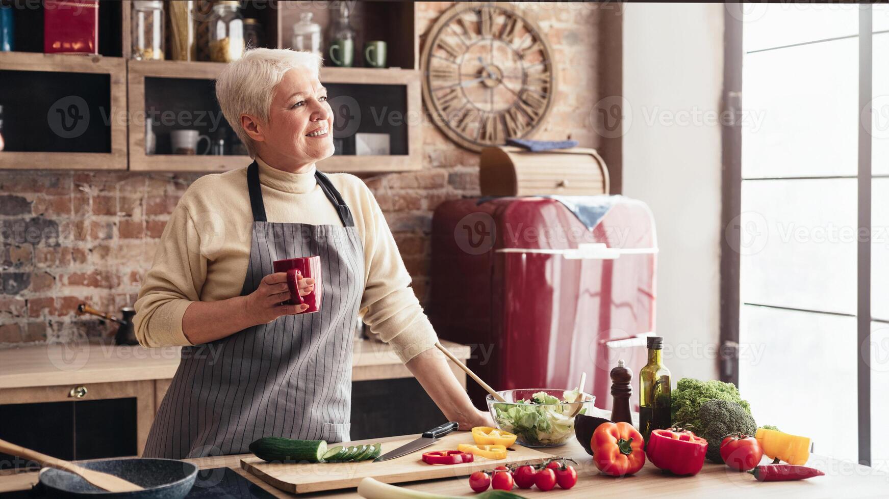 A senior woman stands in her kitchen, preparing a fresh salad. She holds a mug in one hand and stirs a bowl of salad with the other. The kitchen is modern and well-lit with a large window photo