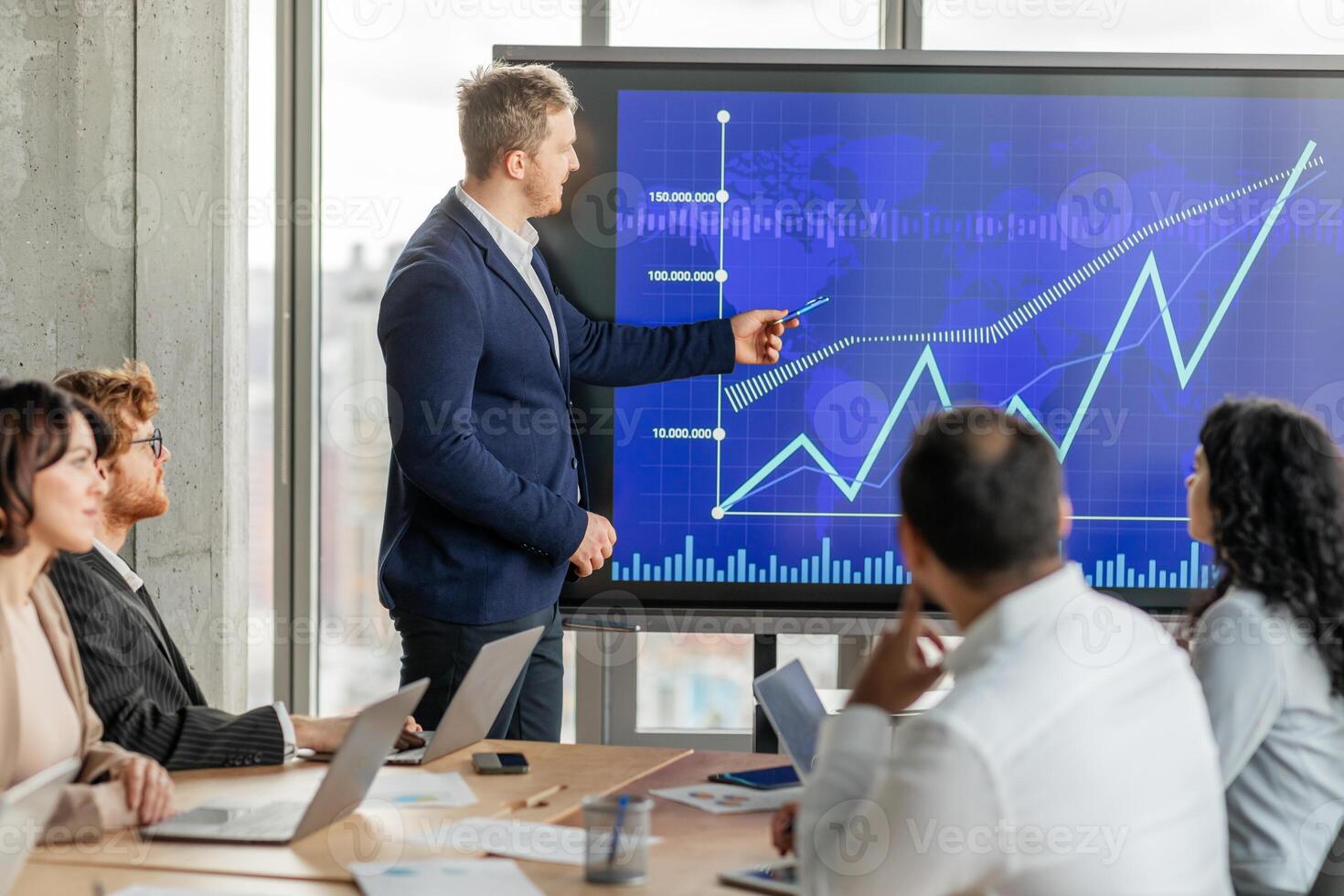 A businessman in a suit points to an upward trending line graph on a large screen while presenting to his team during a meeting. His team sits around a table with laptops open photo