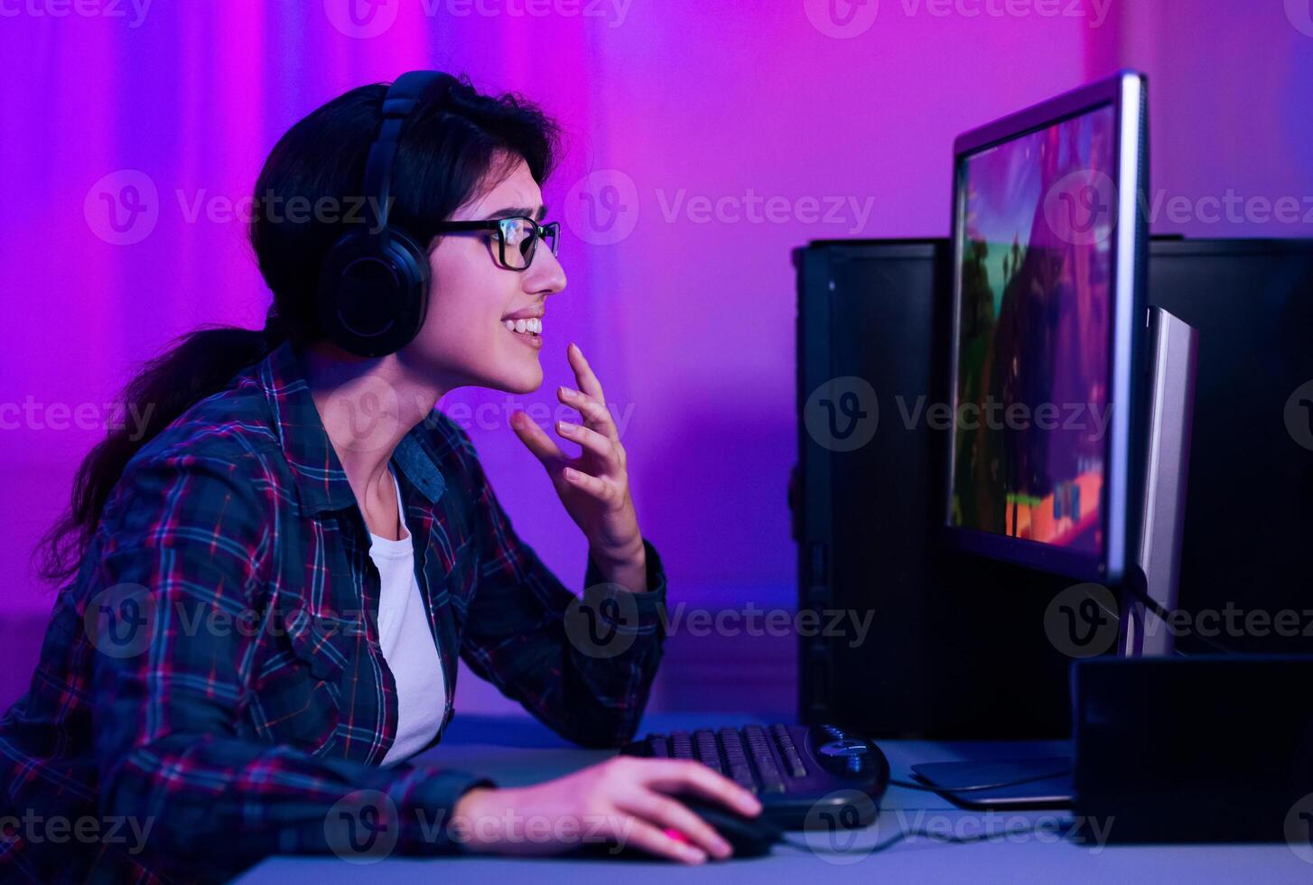 A woman with headphones on is playing a game on a computer. The woman is smiling and has her hand on her chin as she looks intently at the screen. The room is dimly lit with purple lights. photo