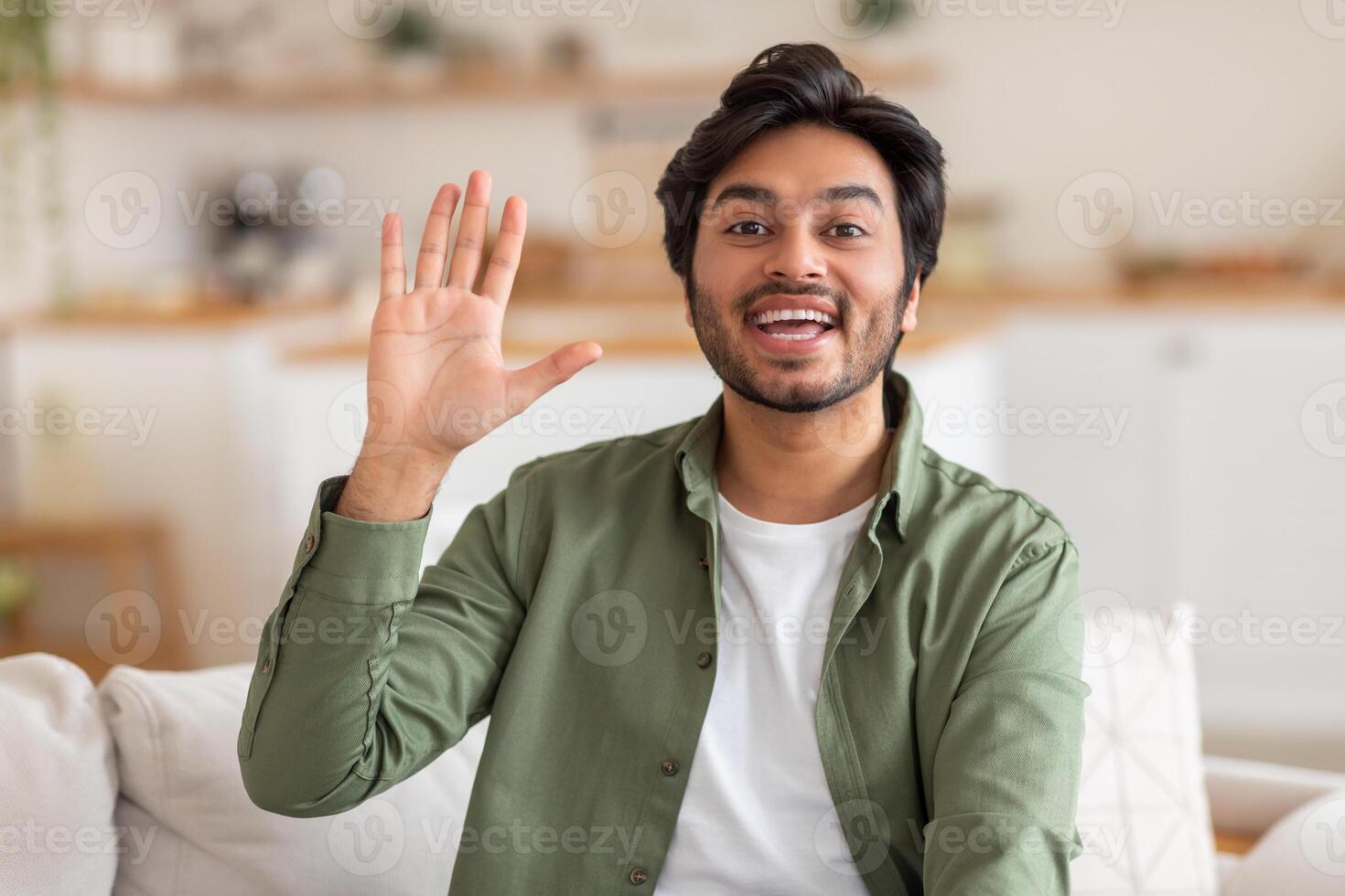 Arab man with dark hair and a beard sits on a white couch in a home. He smiles and waves his right hand, greeting someone. The man is wearing a green button-up shirt over a white t-shirt. photo