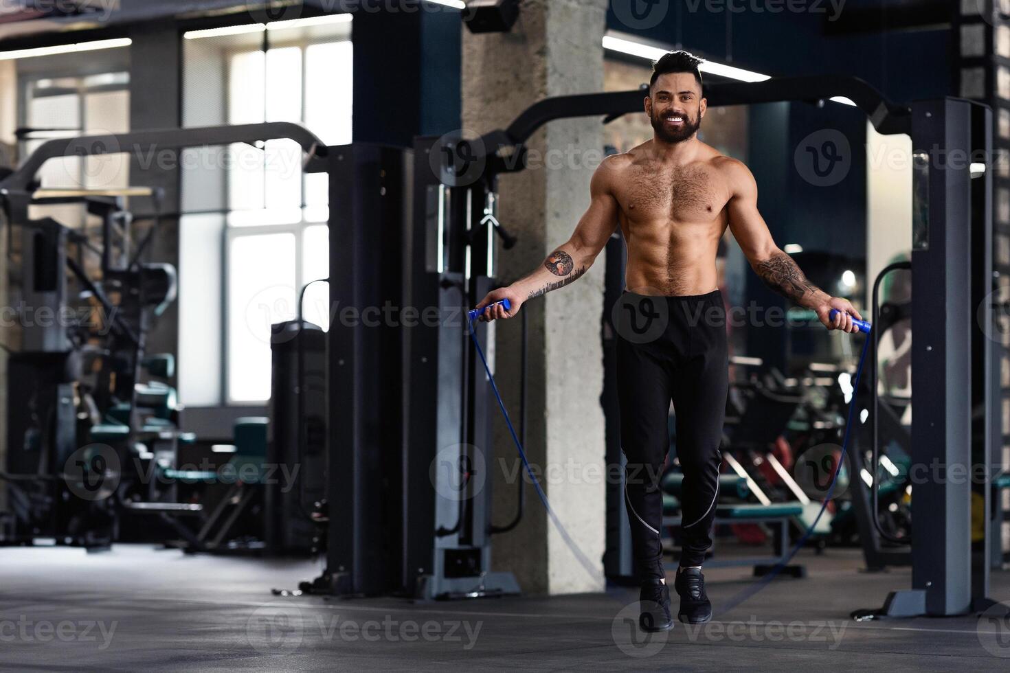 A shirtless man with tattoos is jumping rope in a gym. He is smiling and appears to be enjoying his workout. photo