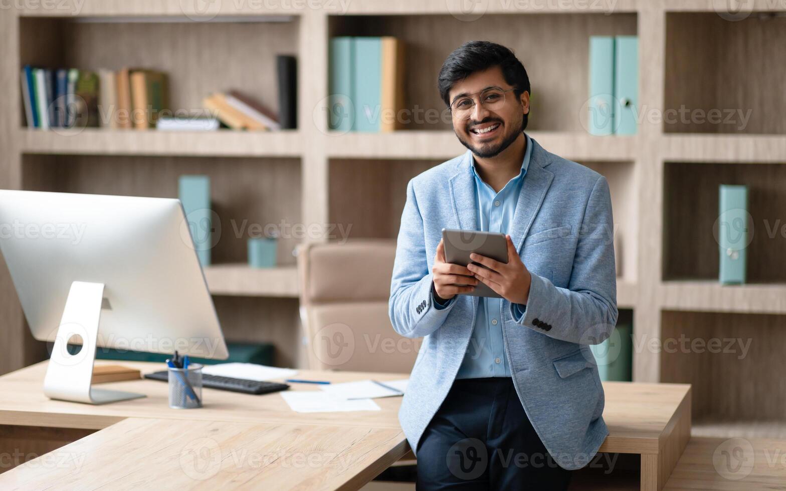 A smiling Indian businessman wearing a blue blazer stands in a modern office, leaning against a desk with a computer monitor in the background. He is holding a tablet in his hands photo