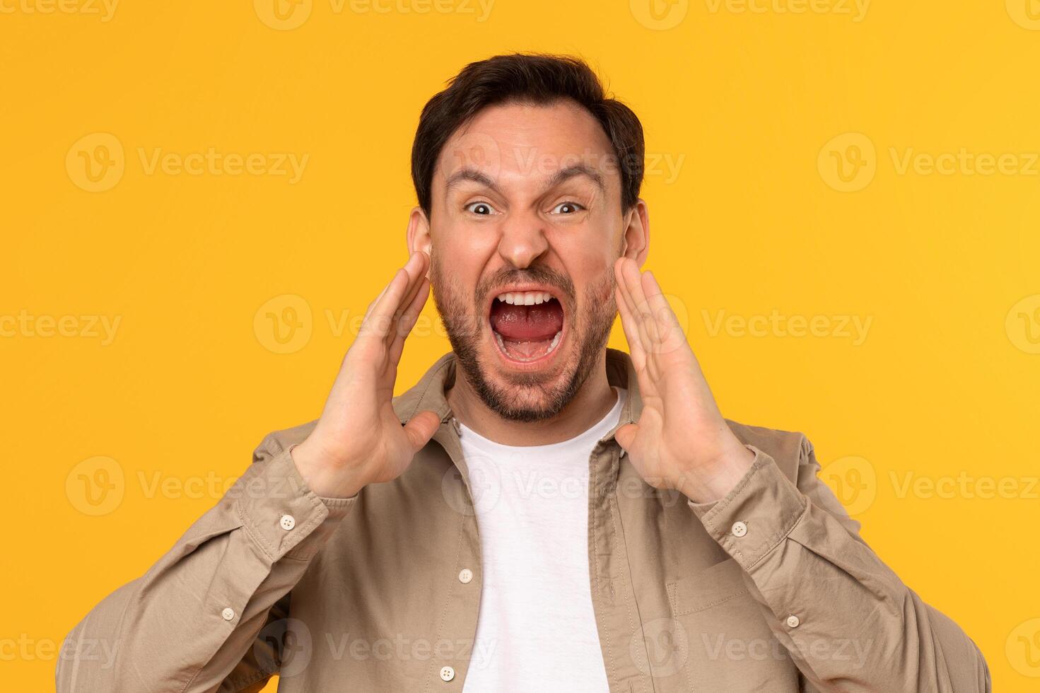 A man with brown hair and a beard is shouting with his mouth open and his hands cupped. He is wearing light brown button-down shirt over white t-shirt. He is standing in front of yellow background. photo