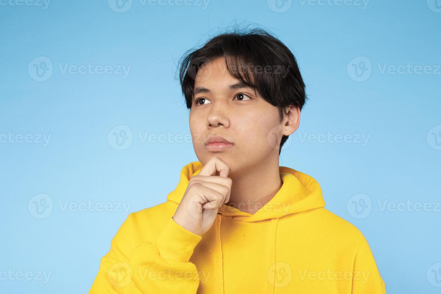 Asian young guy dressed in a bright yellow hoodie stands against a solid blue background, appearing deep in thought. He holds his hand to his chin, looking upwards. photo