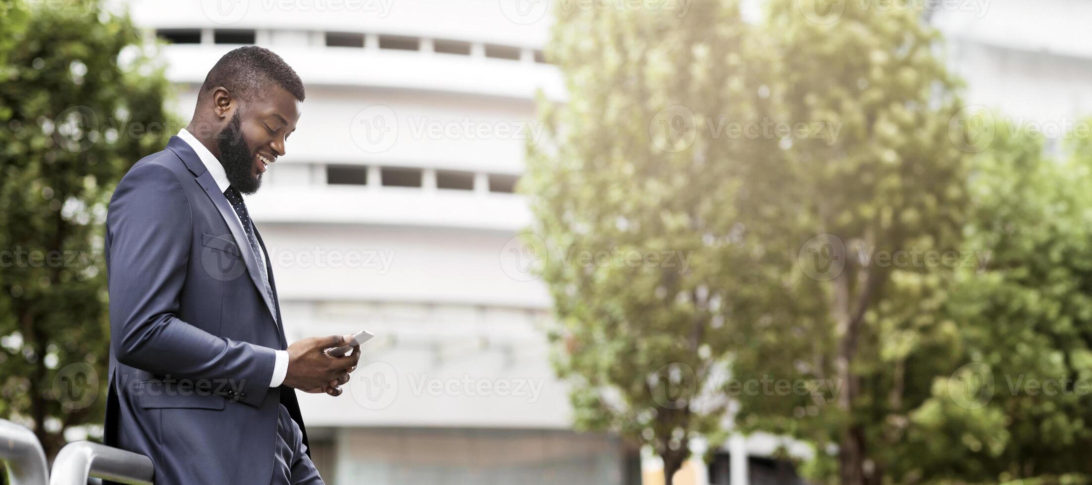 Attractive happy businessman is texting on cell phone near modern office building, side view with copy space. Business, technology and communication concept photo