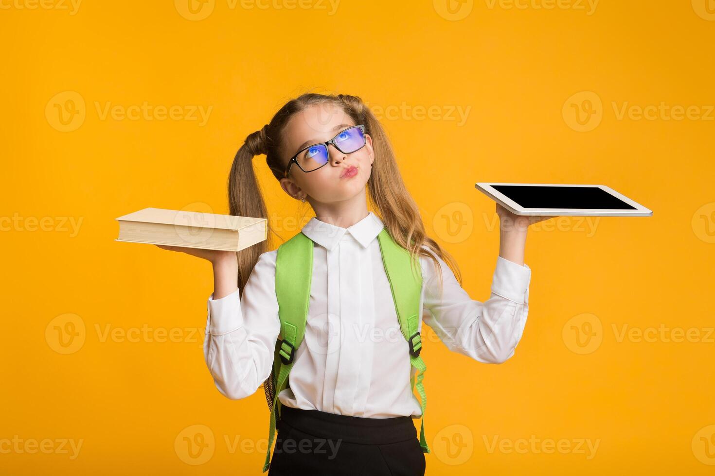 Books Or Gadgets. Nurdy School Girl Comparing Schoolbook And Digital Tablet Over Yellow Background. Empty Space photo