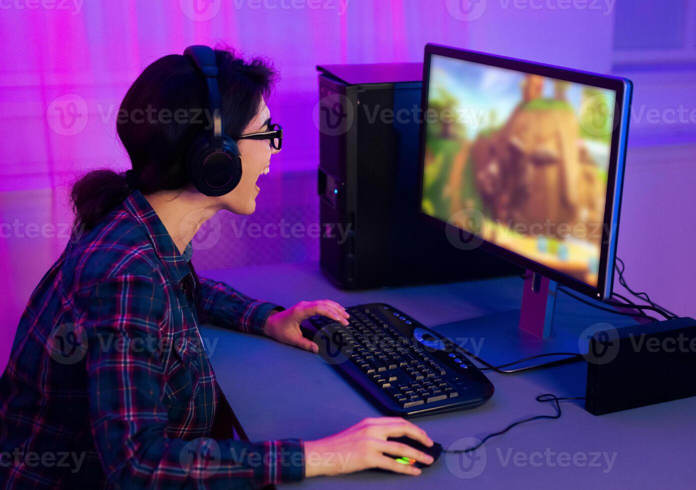 This image shows a young woman wearing a plaid shirt and headphones, playing a game on a computer. She is sitting at a desk with a keyboard and mouse. photo