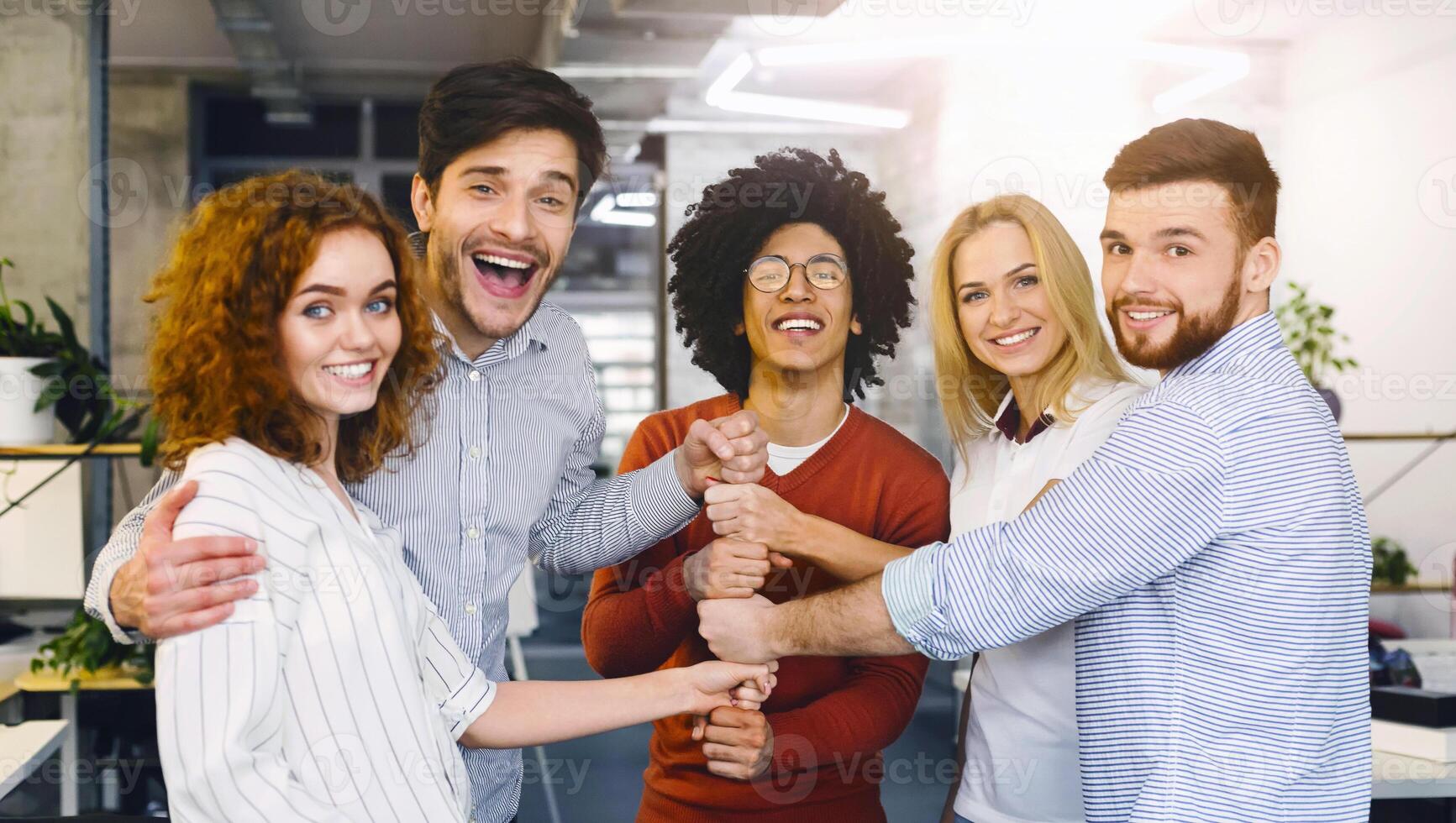 A group of five joyful colleagues forms a huddle, sharing a moment of success with bright smiles and interlocking hands in a gesture of unity and teamwork. The modern office setting photo