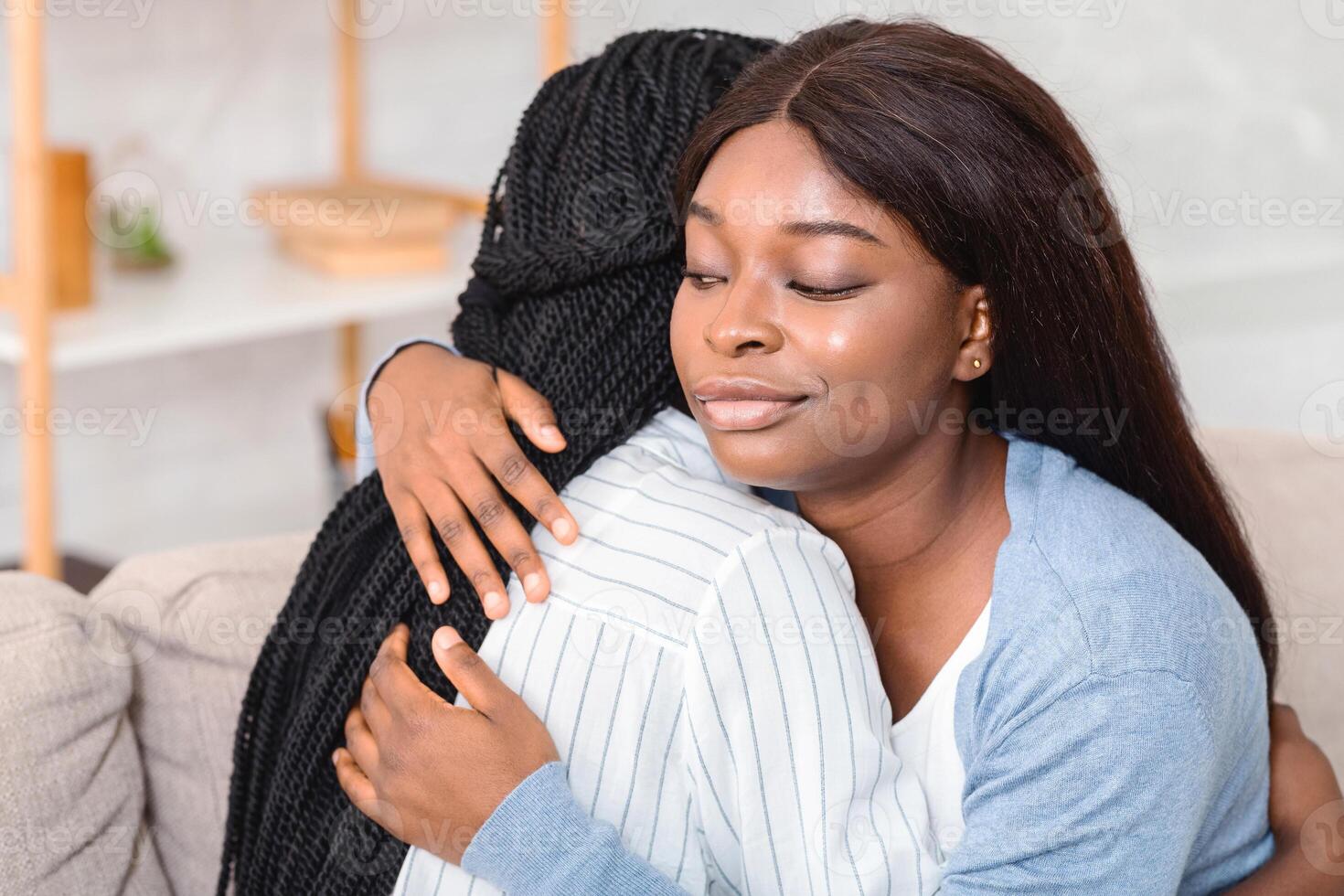 Hypocrisy In Friendship Concept. Afro Girl Hugging Upset Friend With Sneaky Face Expression, Sitting On Couch At Home, Closeup photo