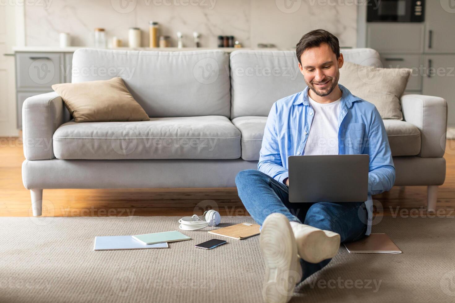 A man is seated on the floor with a laptop open in front of him. He appears focused on the screen, typing and interacting with the device. The room is dimly lit, and there are no other people present. photo