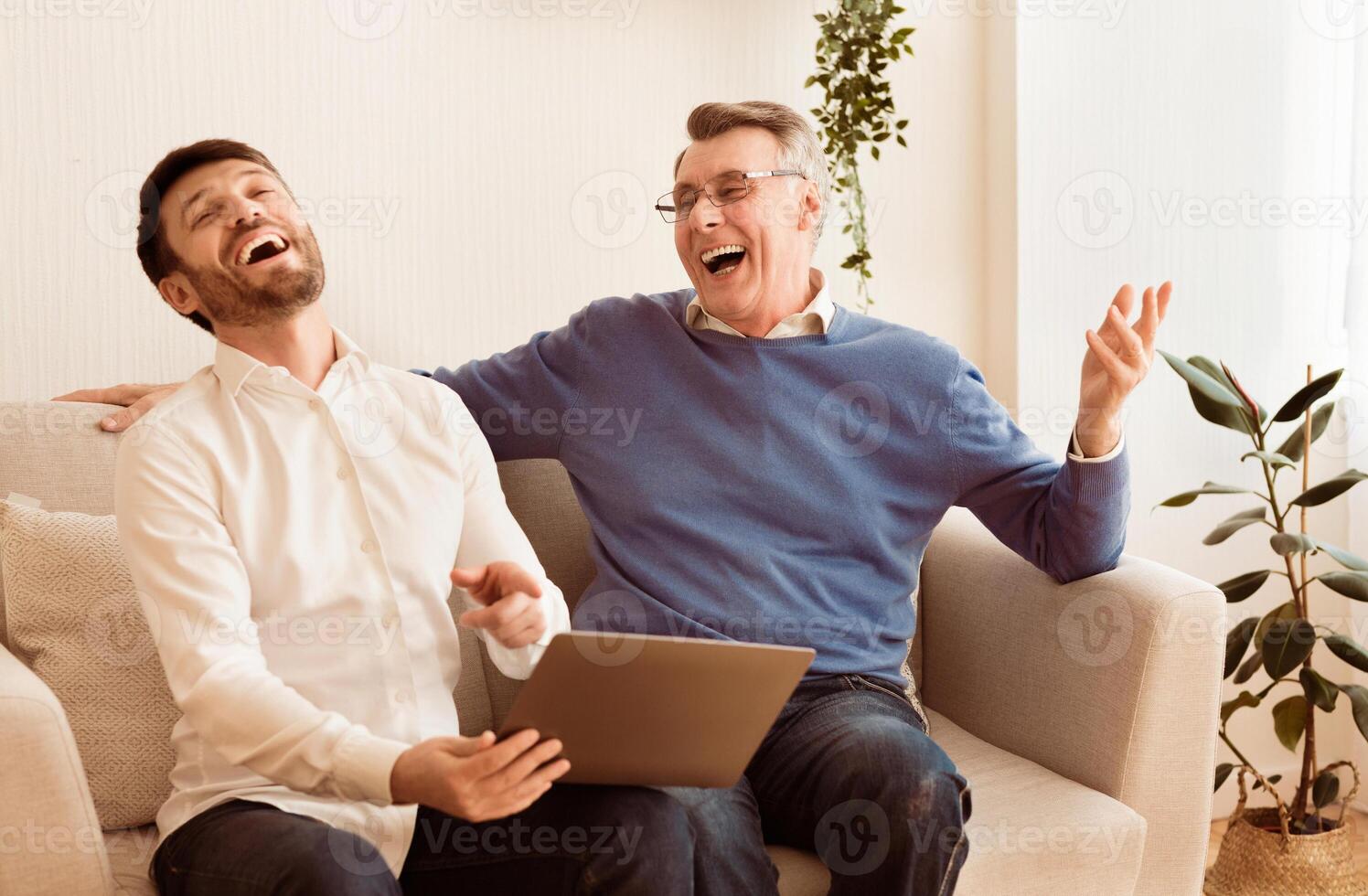 Cheerful Middle-Aged Son And Senior Father Using Laptop And Laughing Sitting On Couch At Home. Selective Focus photo