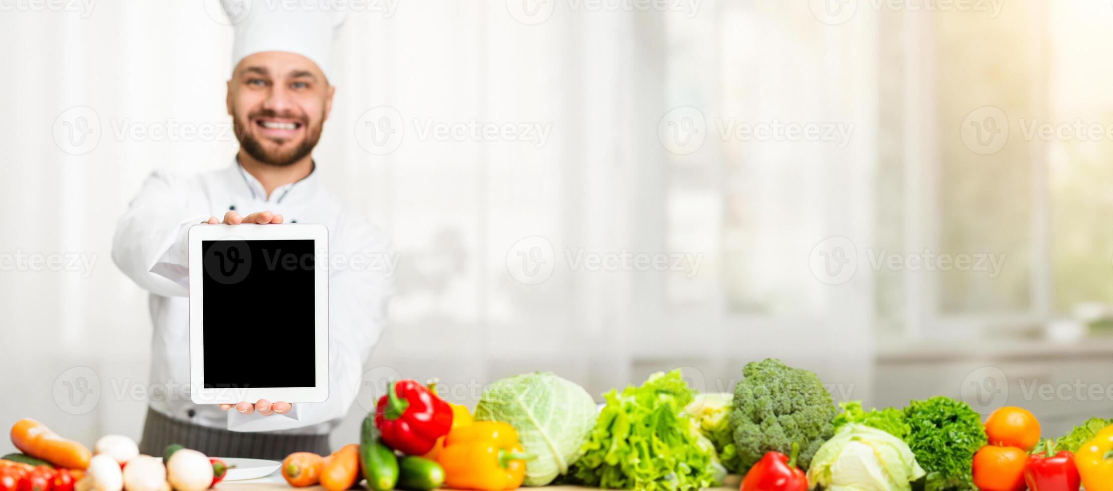 Joyful Chef Guy Showing Digital Tablet With Blank Screen To Camera Standing In Restaurant Kitchen Indoor. Shallow Depth, Mockup photo