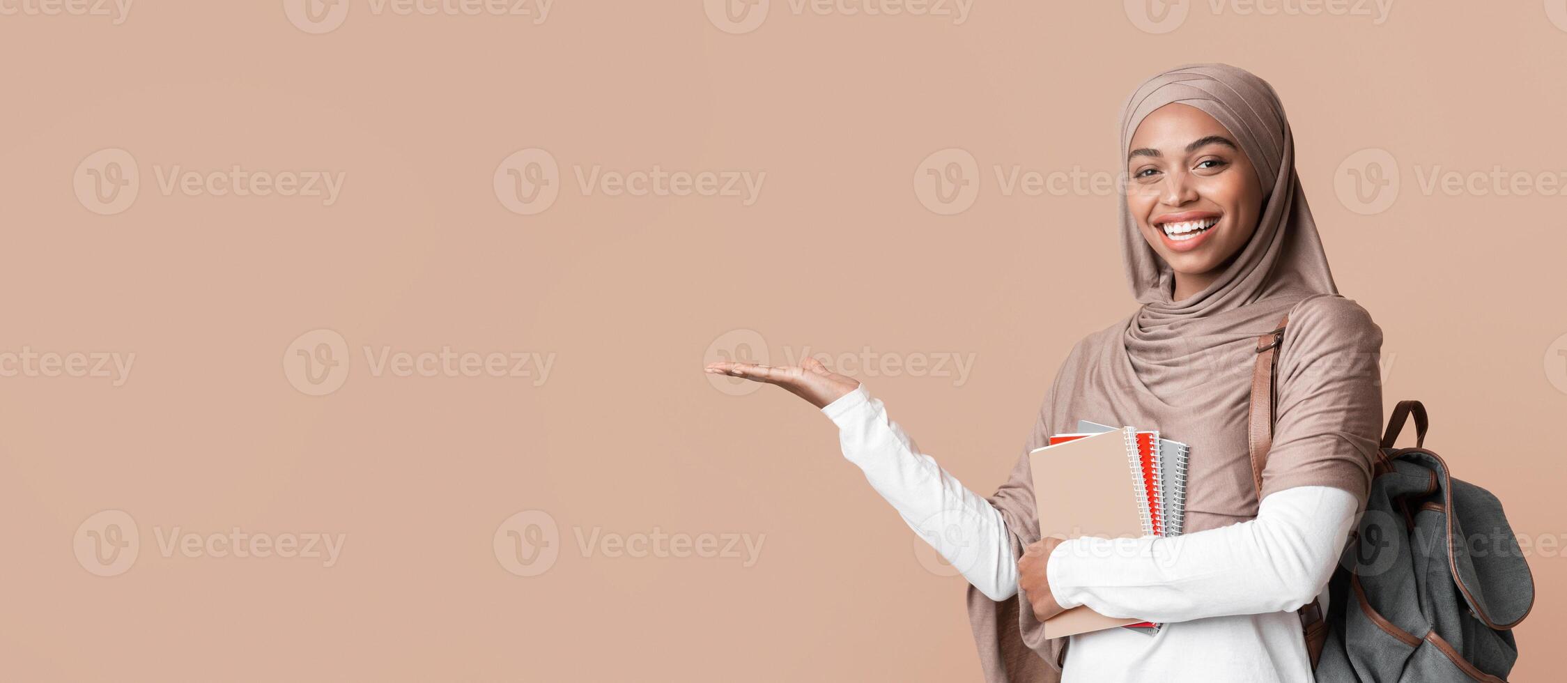 Cheerful black islamic female student in hijab pointing aside at copy space on yellow background with hand or holding invisible object on her palm photo
