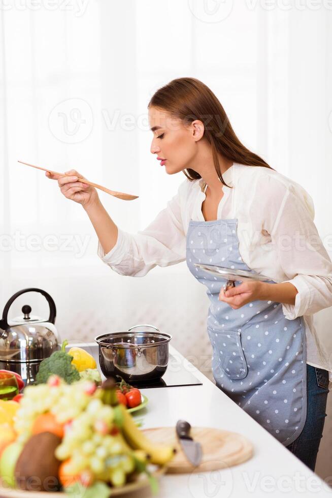 A woman in a blue apron is standing in her kitchen, tasting soup with a wooden spoon. She has a pot on the stove and fresh ingredients on the counter. photo