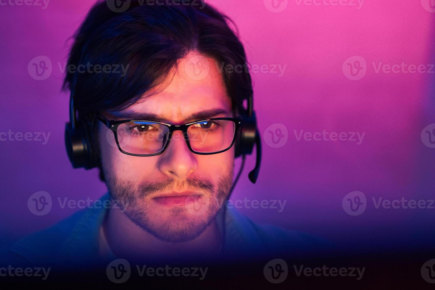 This image shows a young man with dark hair wearing glasses and headphones while looking at a computer screen. He has a serious expression on his face, suggesting he is focused on his task. photo