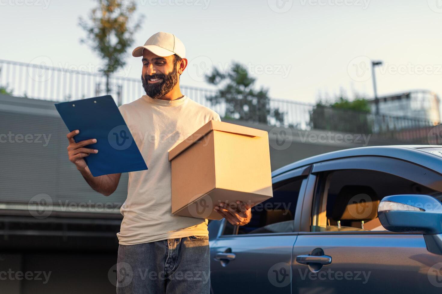 Indian delivery driver is standing next to a blue car in a sunny parking lot. He is holding a large cardboard box in one hand and a clipboard in the other, appearing to check delivery details. photo