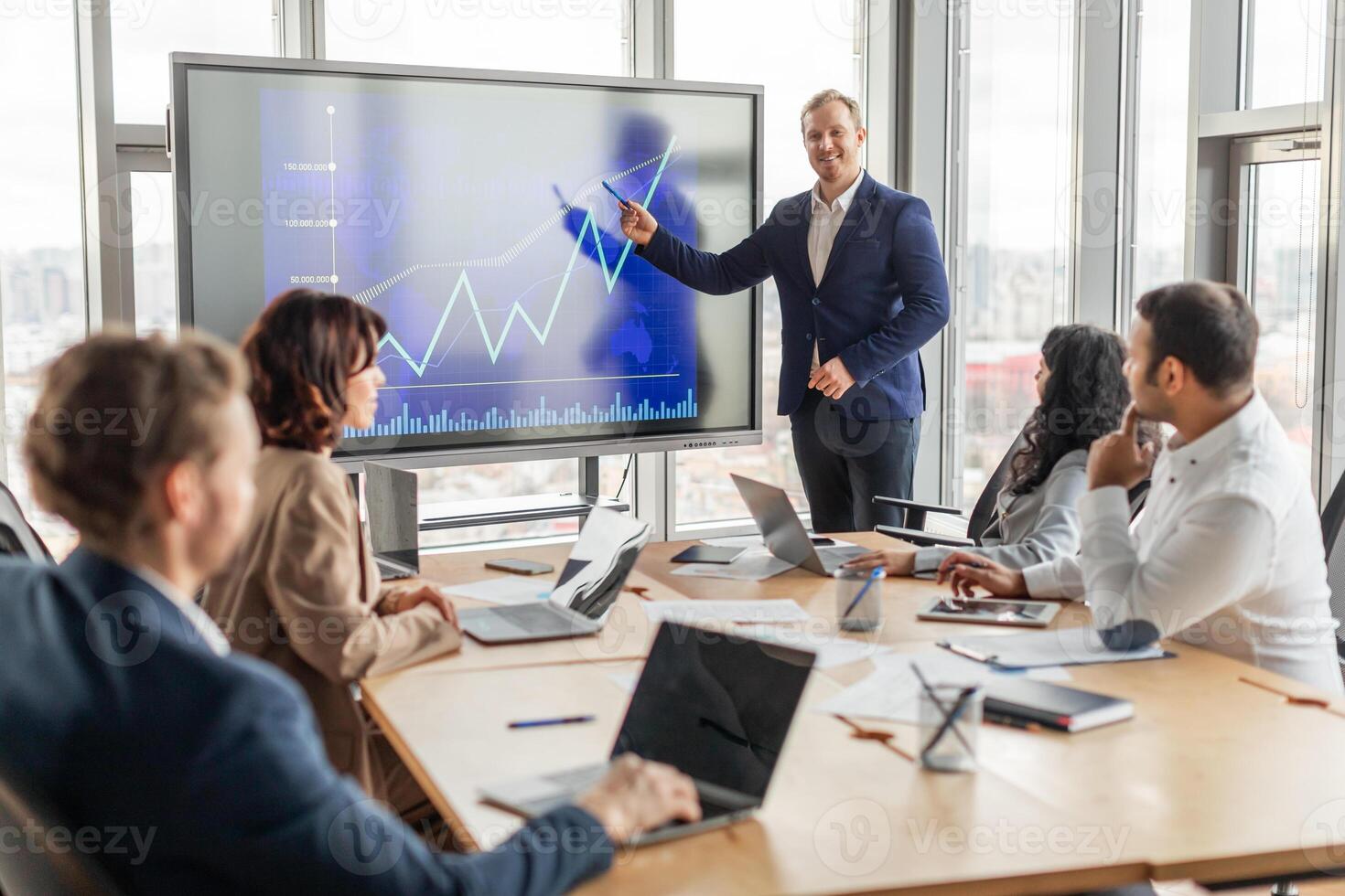 A business professional stands near a large digital screen showing an upward trending line graph. The presenter gestures towards the screen as they explain the chart to a group of colleagues photo