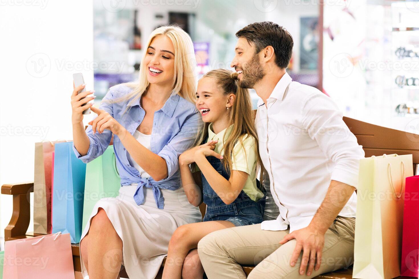 A happy family of three is sitting on a bench in a shopping mall, taking a selfie on a smartphone. The mother is holding the phone, smiling and looking at the camera photo