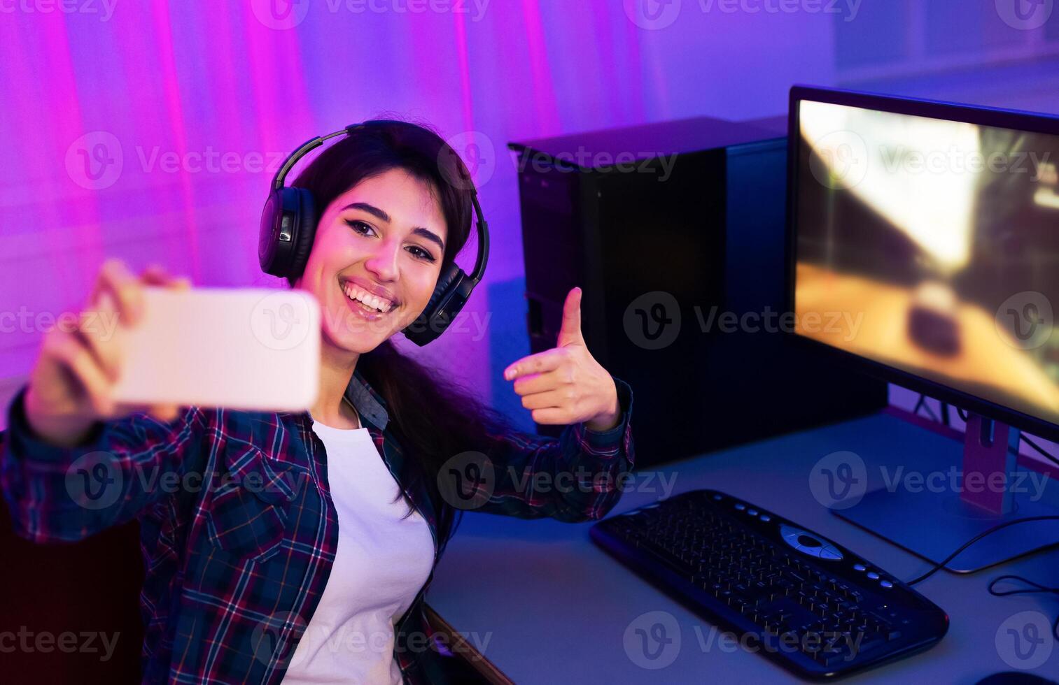 This image shows a woman sitting at a desk with a computer and keyboard. She is wearing headphones and taking a selfie with her smartphone. She is smiling and giving a thumbs-up. photo