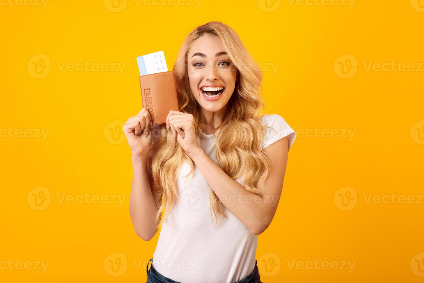 This photo shows a young woman with long blonde hair wearing a white shirt, happily holding a passport and boarding pass in front of a vibrant yellow background.