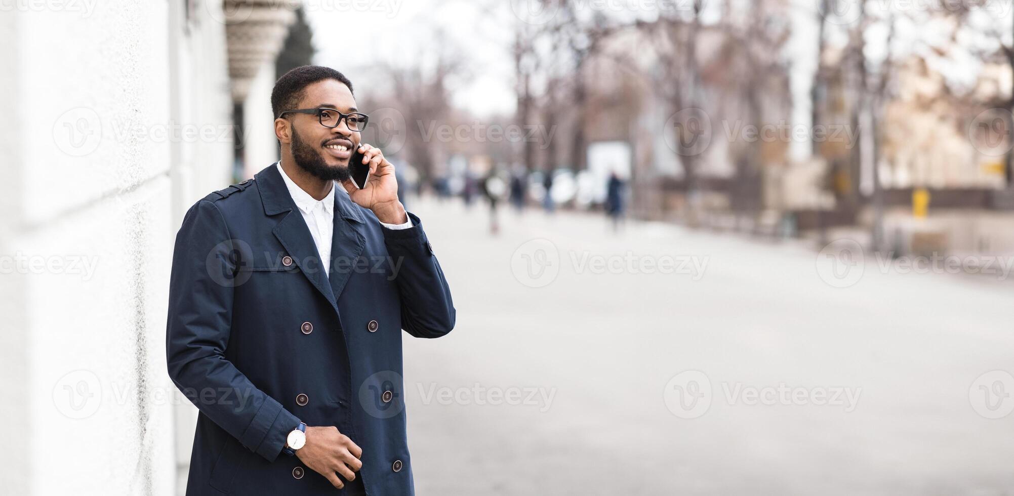African American man in formal outfit have phone conversation outdoors, standing by modern building on the street, panorama with copy space photo