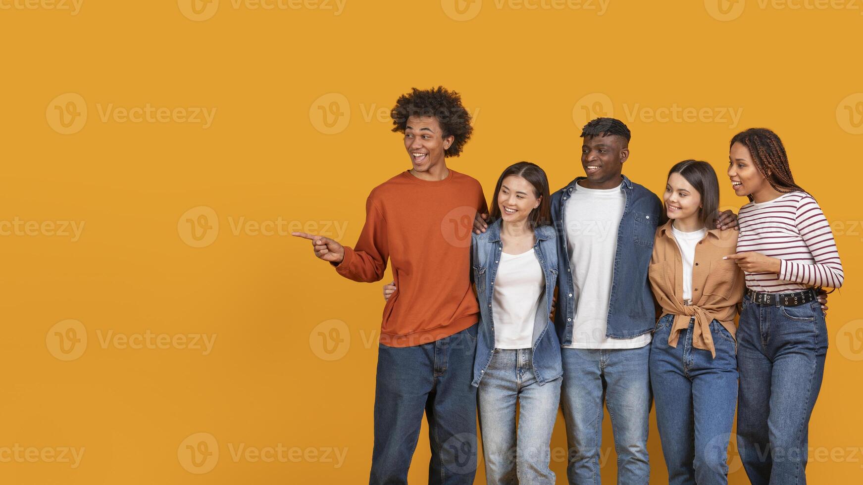 A group of five friends stand together in front of a bright yellow background. They are smiling and looking happy, with one person pointing to the side at copy space photo