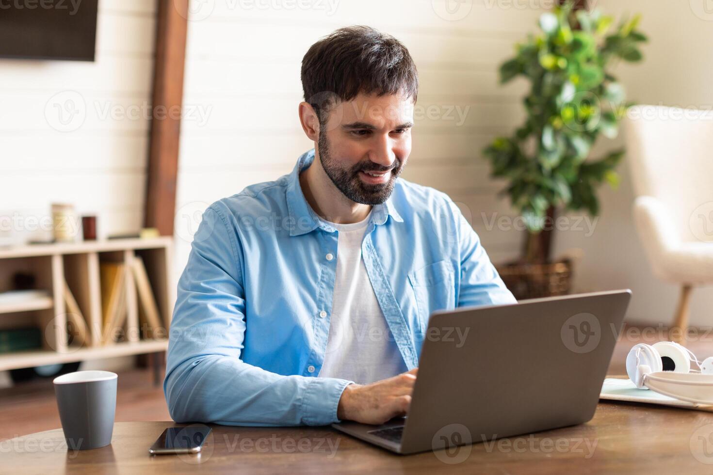 Businessman is seated at a table, focused on his laptop computer screen, typing on the keyboard as he works or browses online. The table is simple, with no distractions, and the background is plain. photo