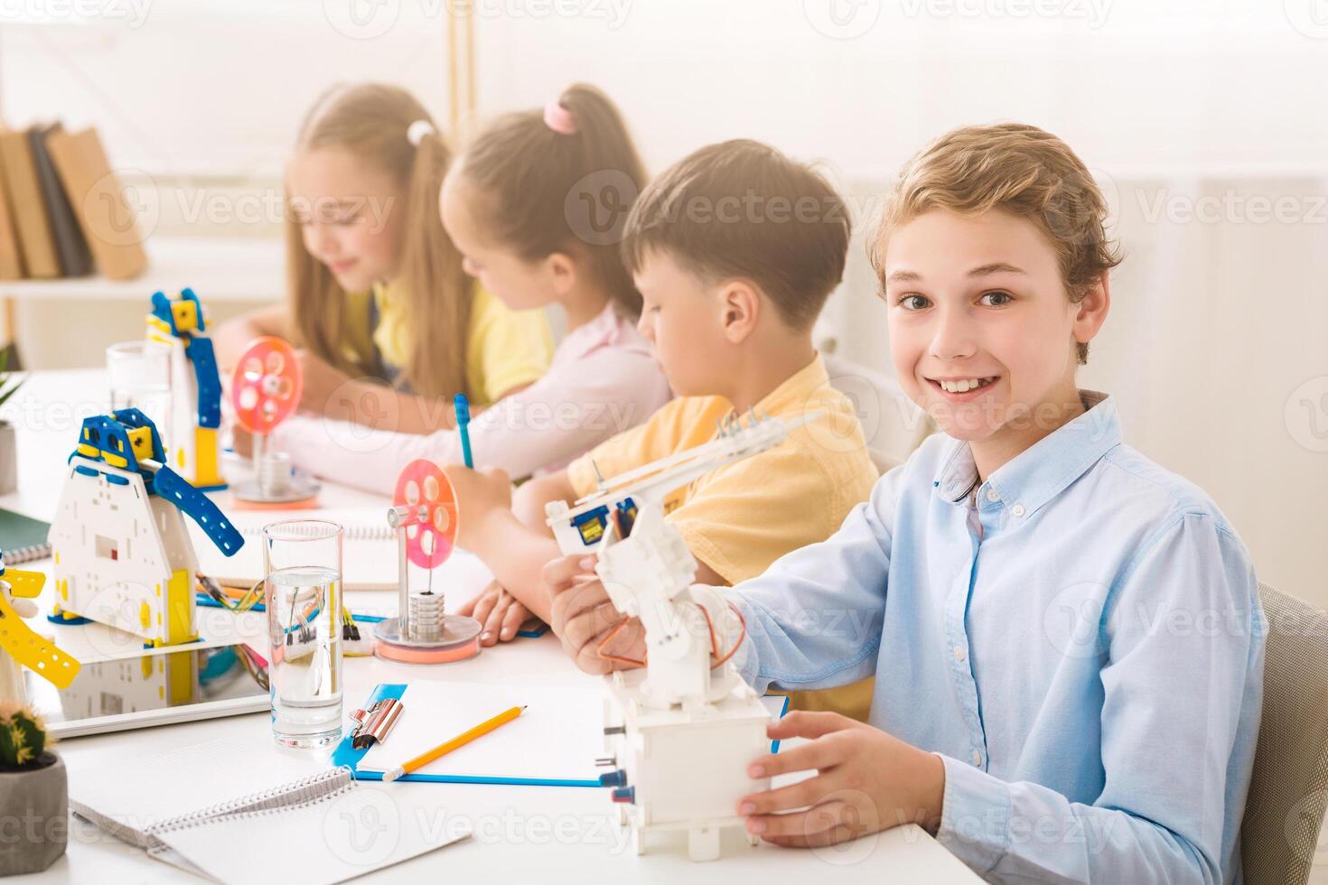 A young boy smiles at the camera while holding a robot during a STEM class. He is sitting at a table with other children who are also working on STEM projects. The classroom is bright and airy photo