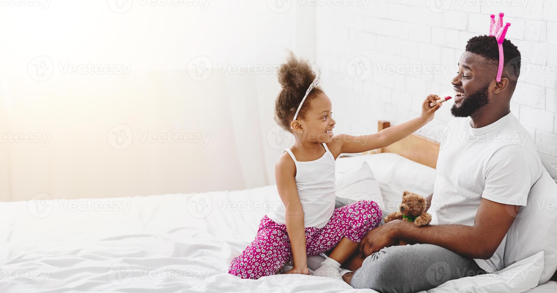 African American little girl, wearing a white tank top and pink floral pants, is applying lipstick to her fathers lips. They are both sitting on a white bed, and the girl is smiling. photo