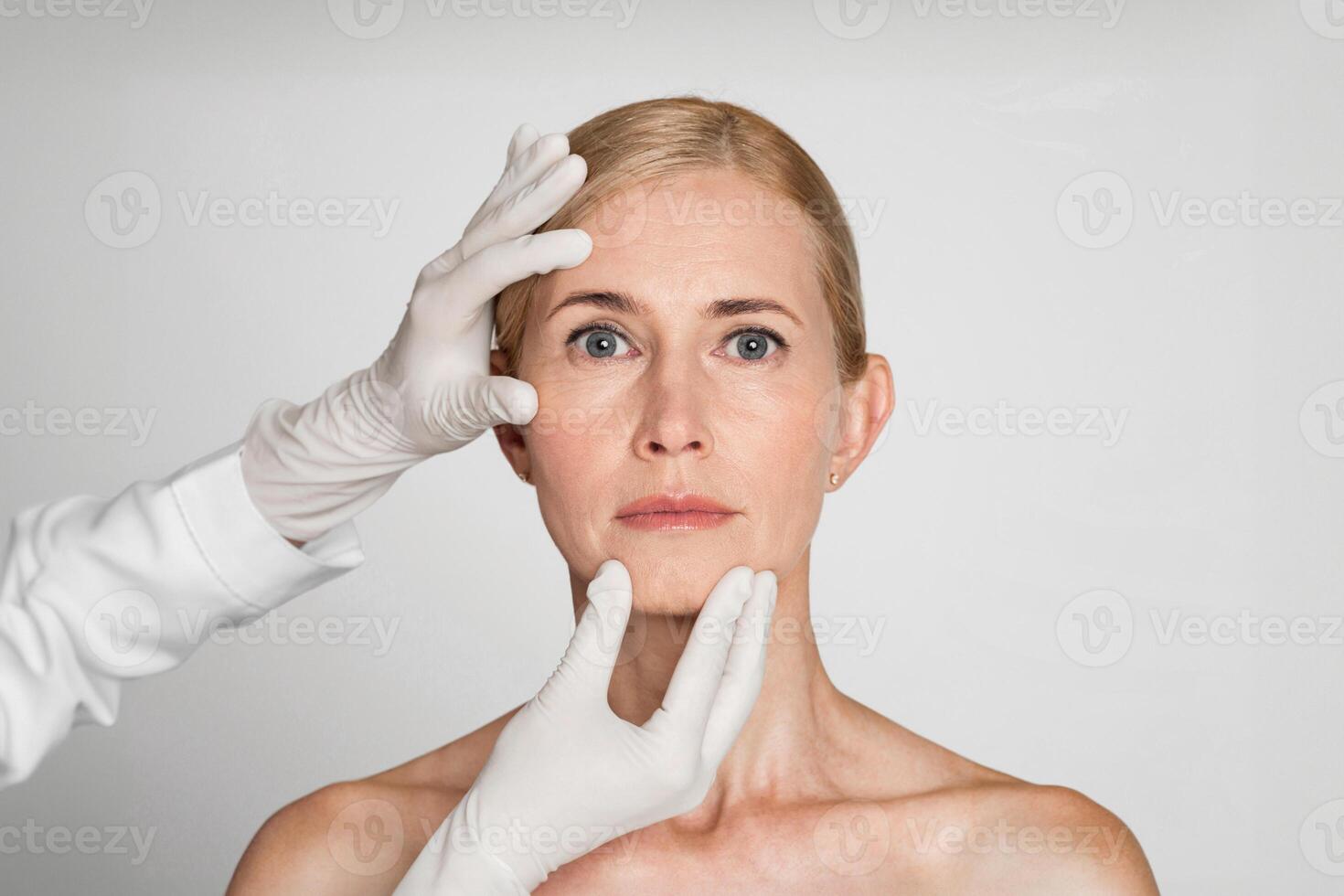 A woman with blonde hair and a serious expression is receiving a facial examination. Two hands, wearing white gloves, are touching her forehead and chin. photo