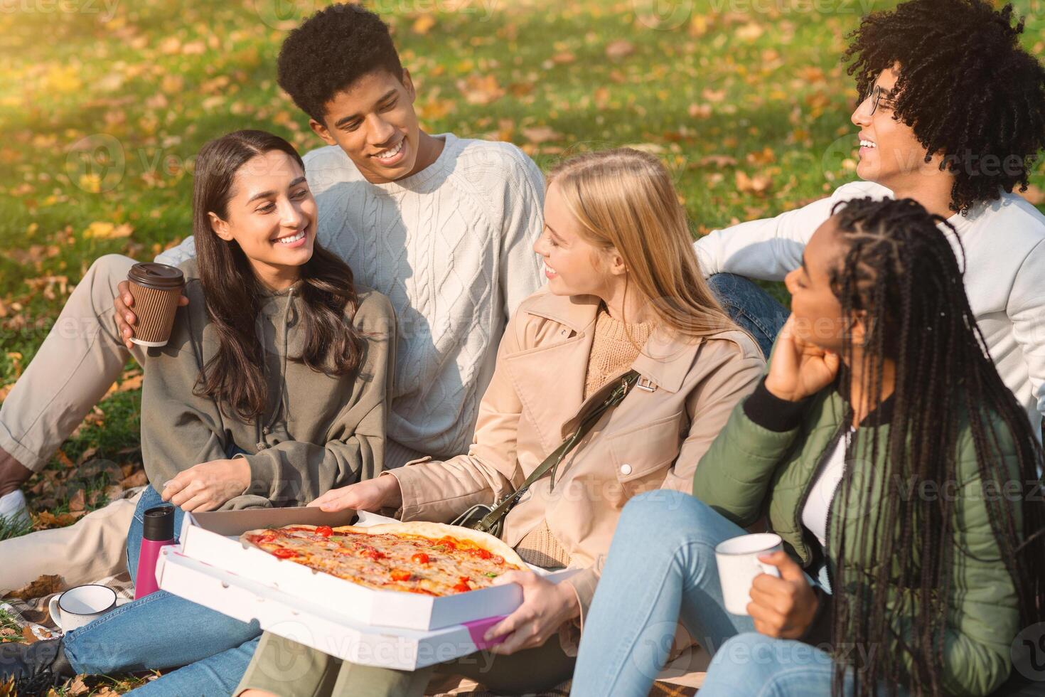 Group of multiethnic teenagers with take away pizza sitting on ground, enjoying weekend together photo