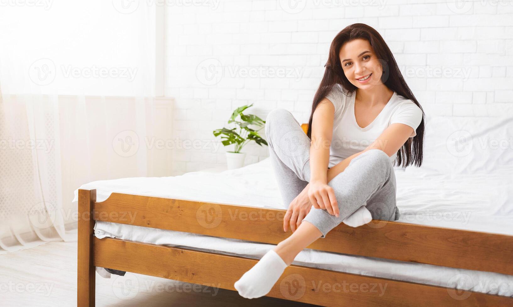 A woman is seated on top of a wooden bed, looking towards the camera. The room is minimally decorated, with the bed being the central focus. photo