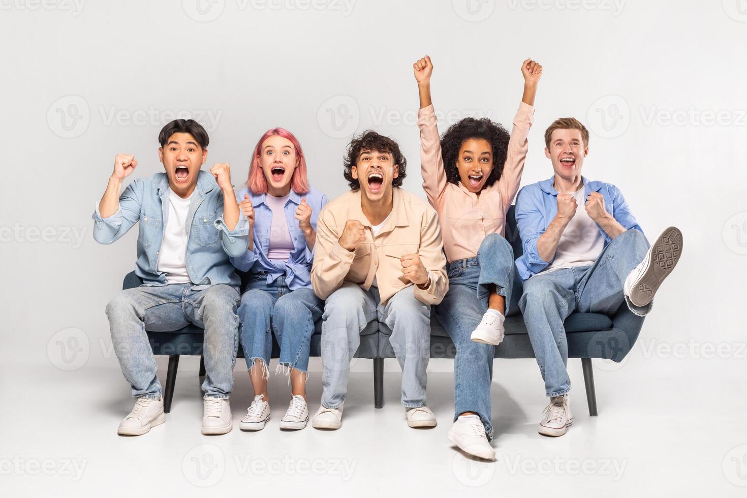 A group of diverse young adults are excitedly celebrating together on a couch. They are all smiling and laughing, with their arms raised in the air. photo