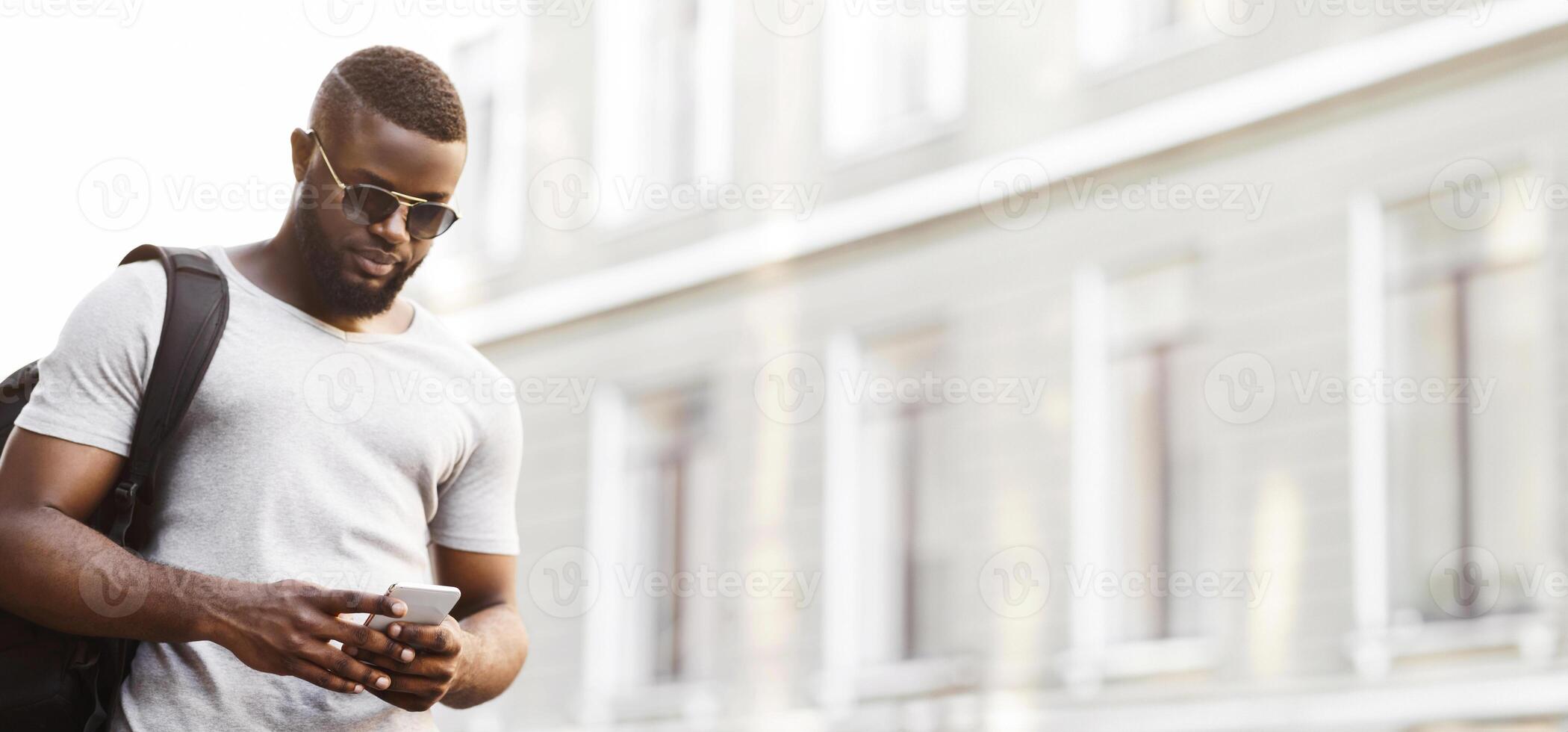 A man wearing sunglasses checks his phone while walking on a city street. photo