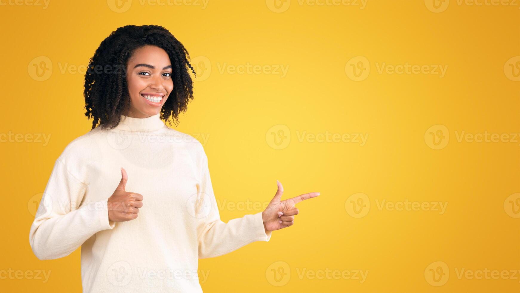 Look There. Happy afro woman pointing index finger at copy space, showing thumb up over yellow studio background photo