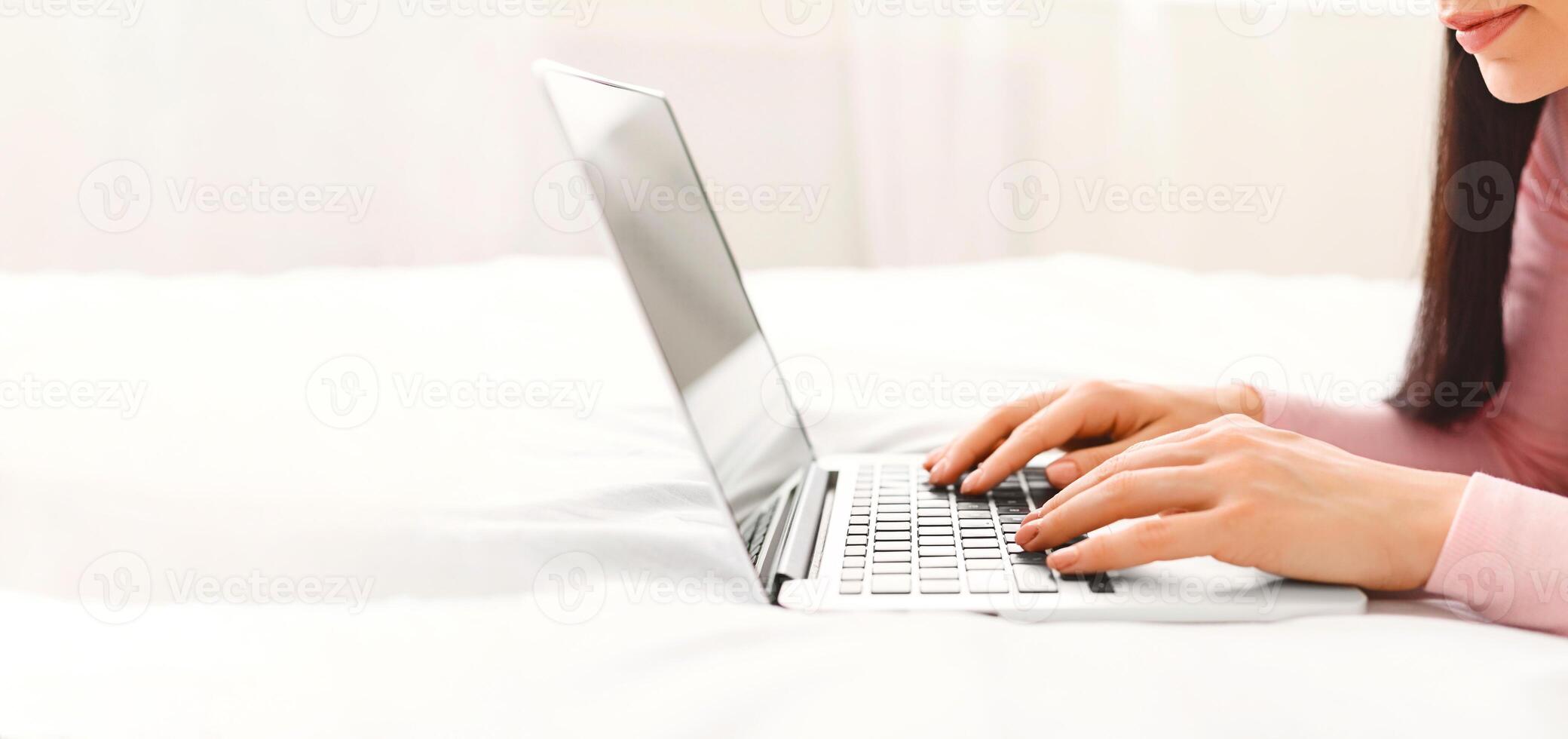 A woman is sitting on a bed, focused on using a laptop computer. She is typing and browsing online content, cropped, panorama photo