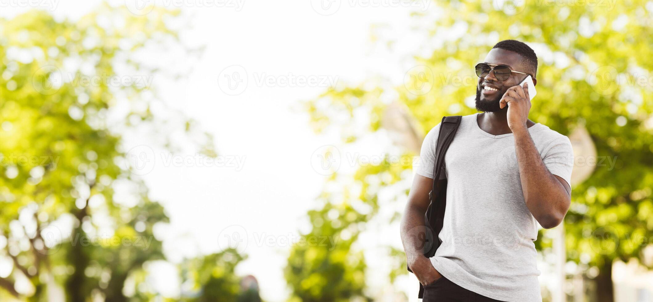 A young man with a backpack smiles while talking on his phone outdoors. He is standing in front of a blurred background of green trees. photo