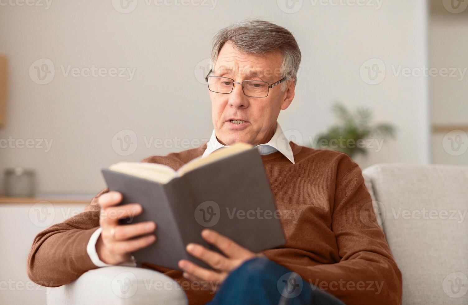 Retired man reading book sitting on couch at home on weekend. Retirement leisure activities. Selective focus photo