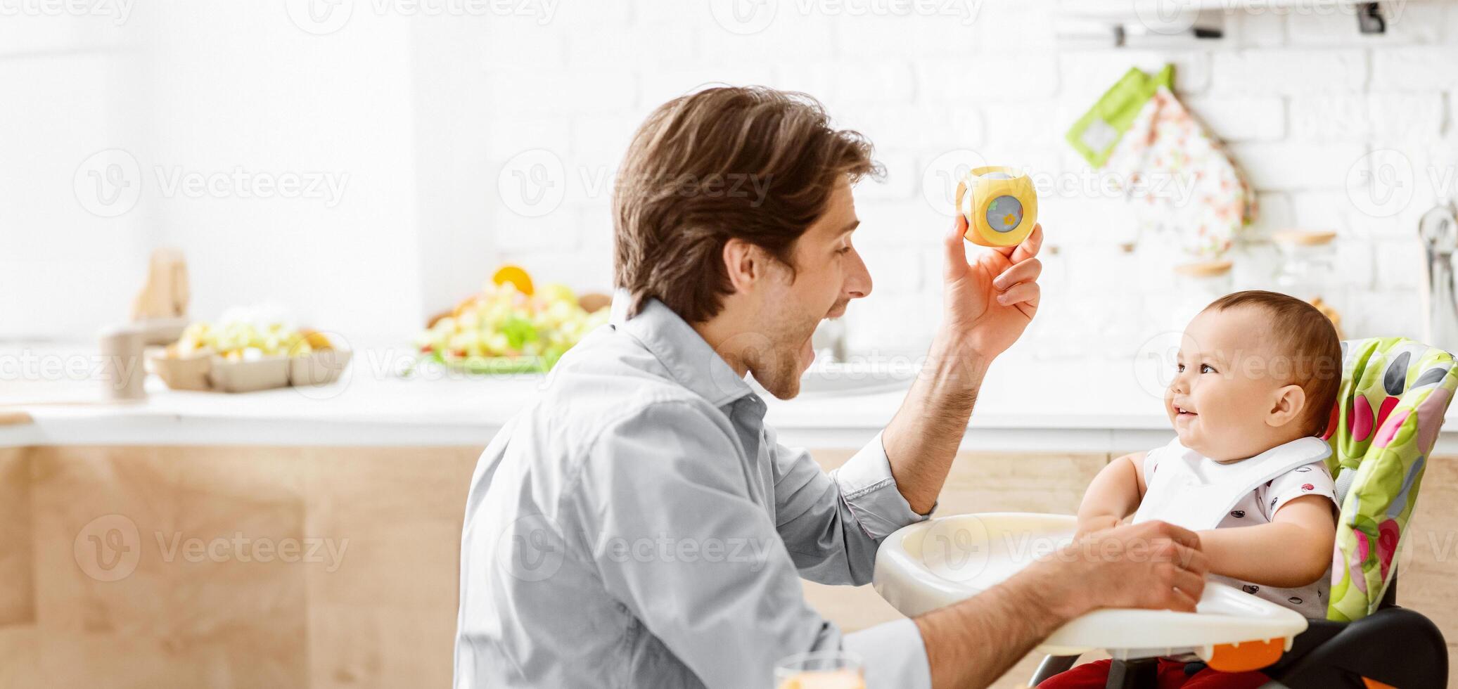 A father sits in a kitchen with a baby in a high chair. He is holding a yellow toy and making the baby laugh. The kitchen is bright and clean. photo
