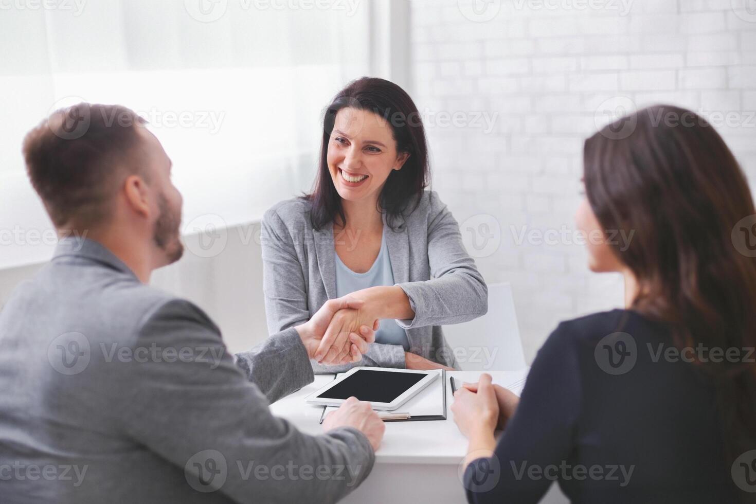A woman in a gray blazer smiles and shakes hands with a man sitting across from her at a table. A third person sits to the woman right and observes the interaction. photo