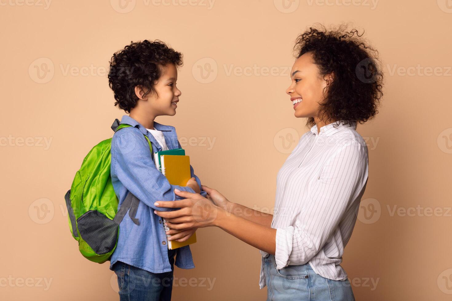 A young African American boy, outfitted with a green backpack and holding colorful books, stands facing his smiling mother as she offers him support and affirmation before he heads off to school photo