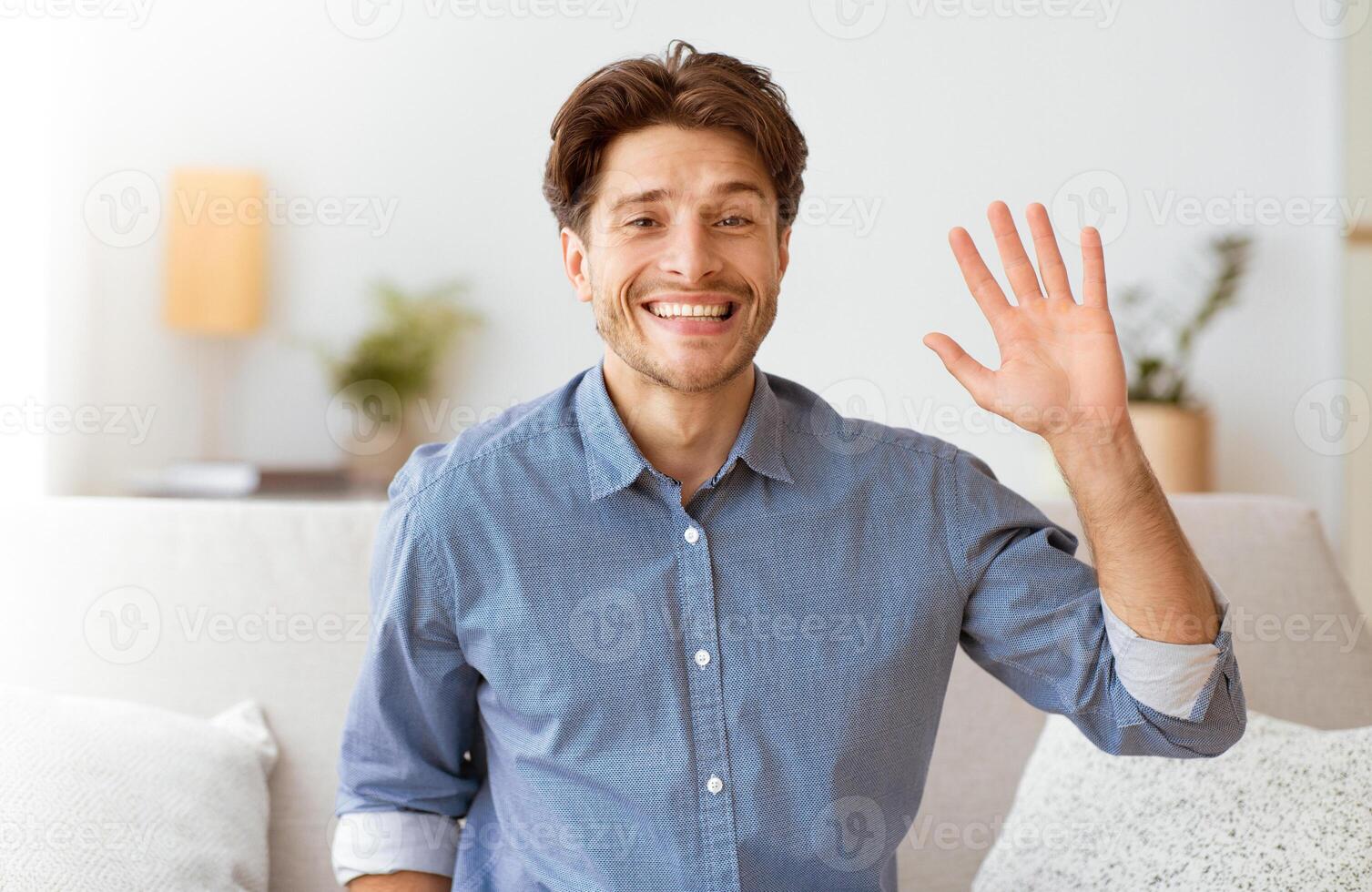A man smiles and waves his hand while sitting on a couch in a living room. photo