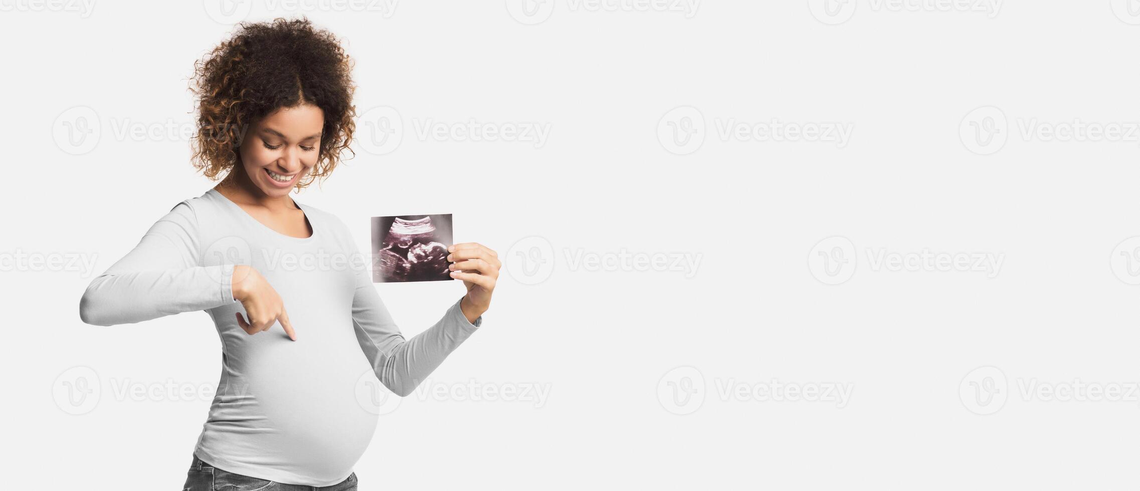 Happy pregnant lady showing ultrasound scans and pointing at belly over white background photo