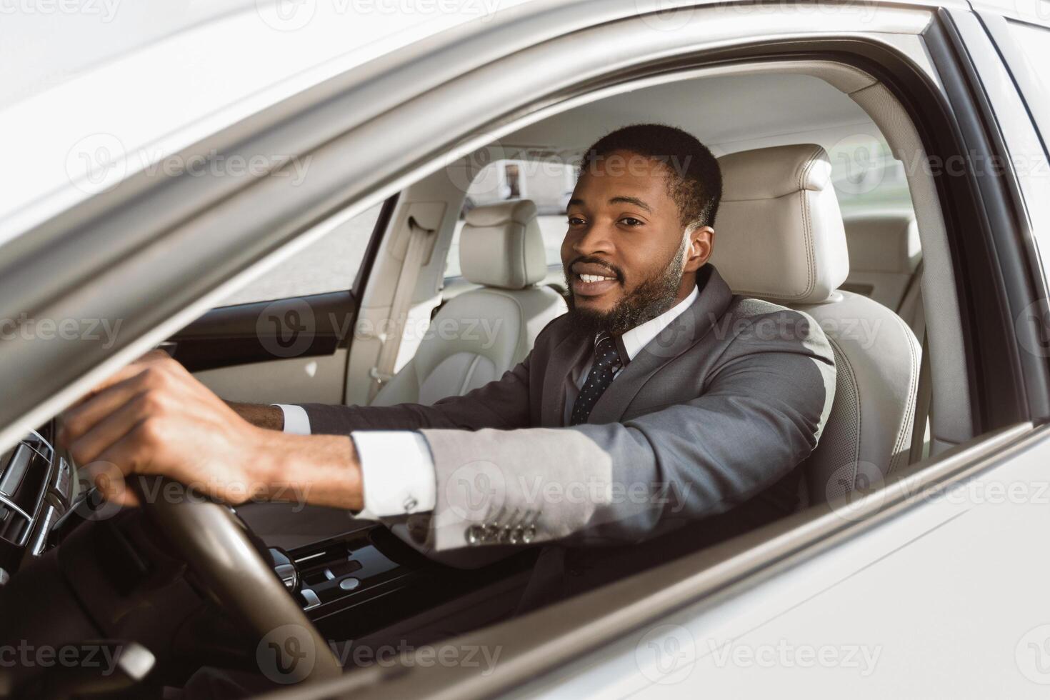 A black man wearing a suit is driving a car on a sunny day. He is smiling and appears to be enjoying the drive. The car is modern and sleek, with a white exterior and a luxurious interior. photo