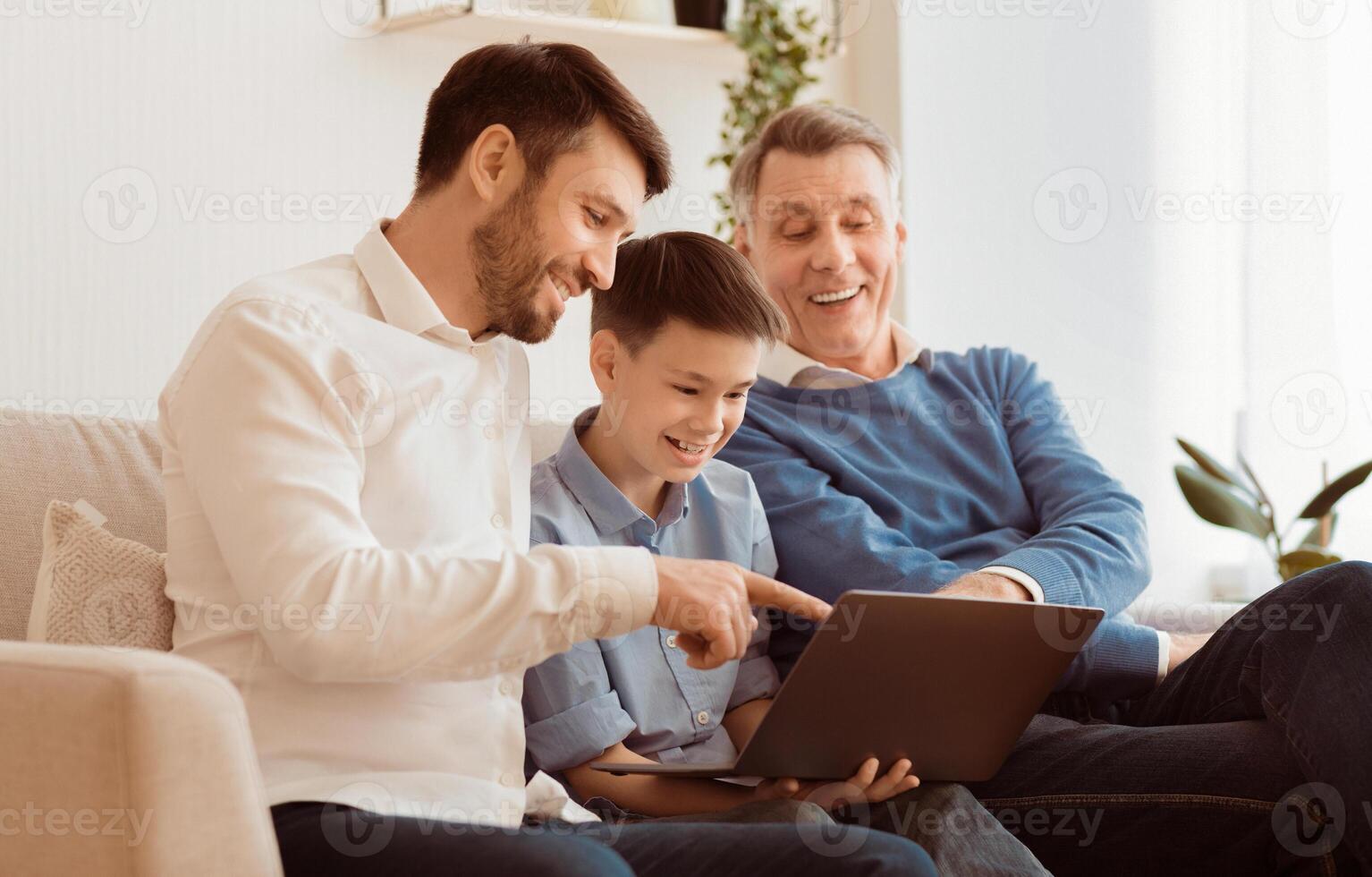 Boy Using Laptop Sitting Between Father And Grandpa Doing Homework Online On Sofa At Home. Selective Focus photo