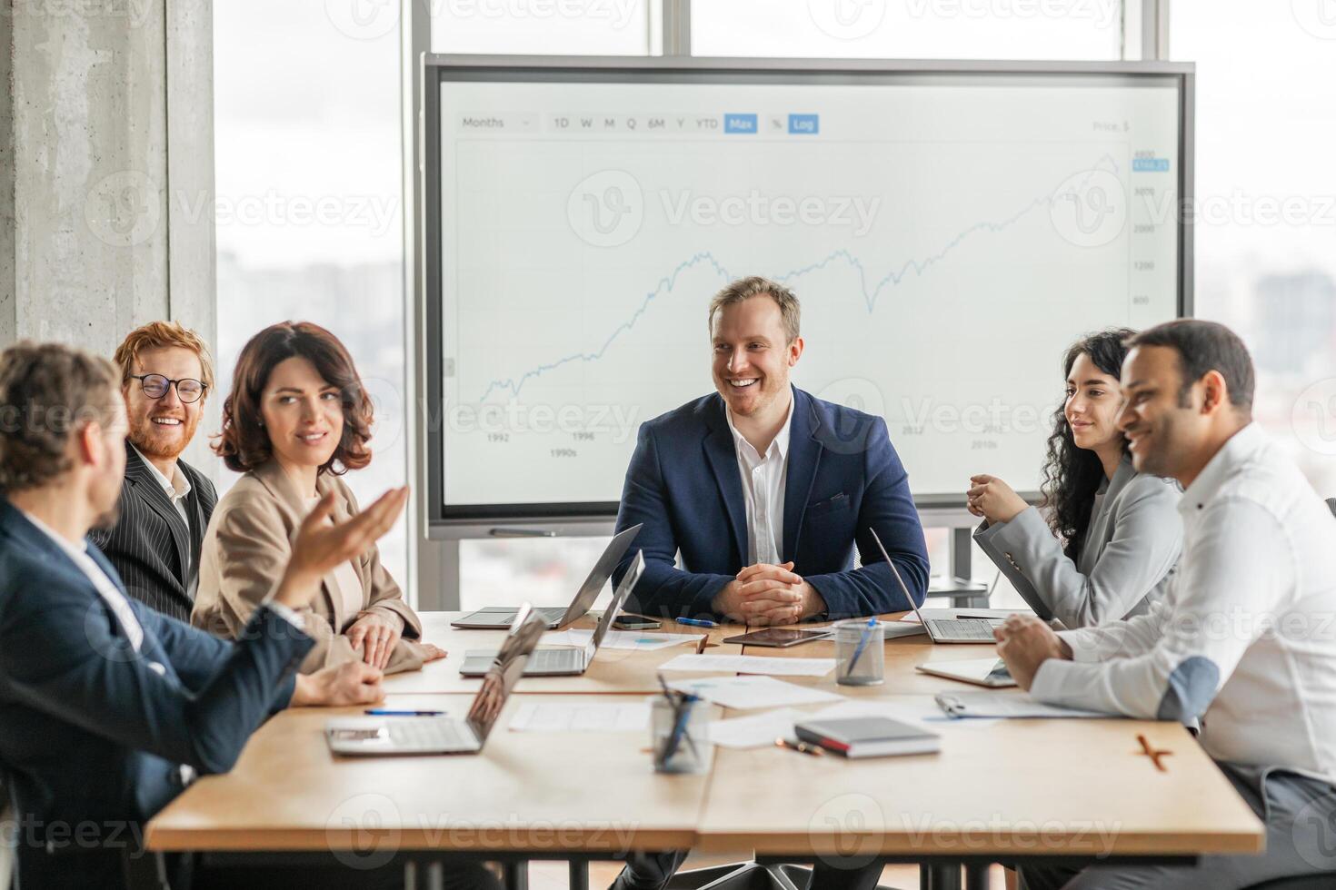 A group of business professionals sit around a large wooden conference table, engaged in a meeting. They are looking at a large screen behind them displaying a projected growth graph photo