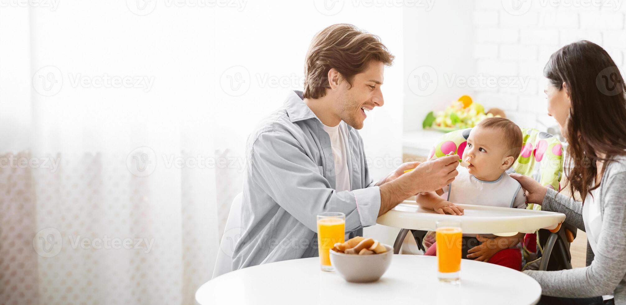A smiling father feeds his baby while his wife watches nearby. They are enjoying a breakfast together at home, with a glass of orange juice and a bowl of snacks on the table, copy space photo