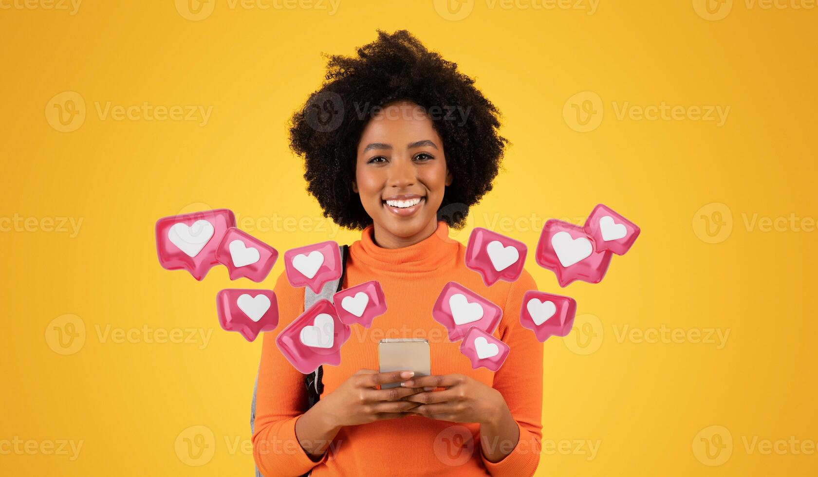 Smiling millennial African American woman with curly hair, wearing an orange turtleneck, texts boyfriend on her phone while carrying a grey backpack on a vibrant yellow background, studio photo