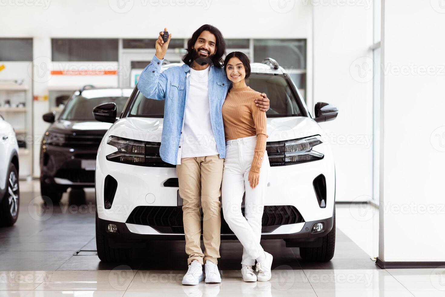 Indian man and woman are standing in front of a car, looking towards the camera. The car is parked on a paved road with trees in the background. photo