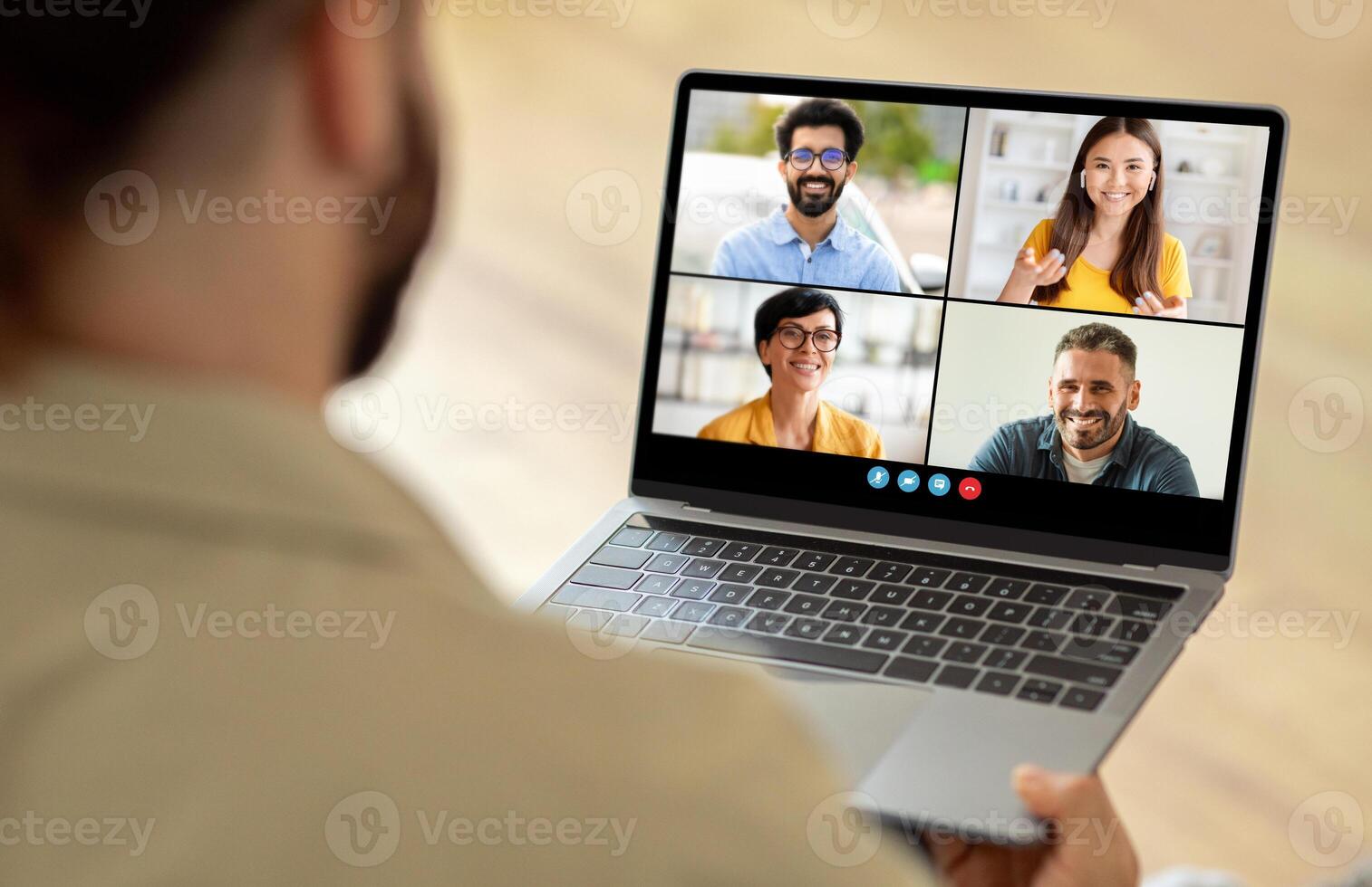 A man sitting at a table is using a laptop, which displays a group of people on the screen. He appears engaged in a virtual meeting or conference with his colleagues. photo